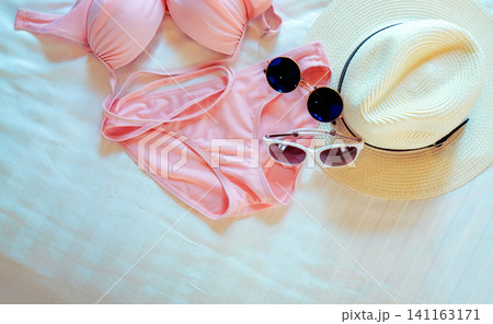 Pink bikini swimwear with stylish sunglasses and a wide brim sun hat arranged on white fabric. Fashionable summer flat lay for tropical vacation, beach holiday, travel, and resort lifestyle concept. 141163171