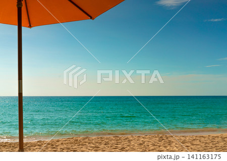 Orange beach umbrella overlooking the turquoise tropical ocean beneath blue sky. Relaxing summer vacation background. Holiday, tourism, luxury resort, summer vacation travel, and wellness concept. Orange beach umbrella overlooking the turquoise tropical ocean beneath blue sky. Relaxing summer vacation background. Holiday, tourism, luxury resort, summer vacation travel, and wellness concept. 141163175