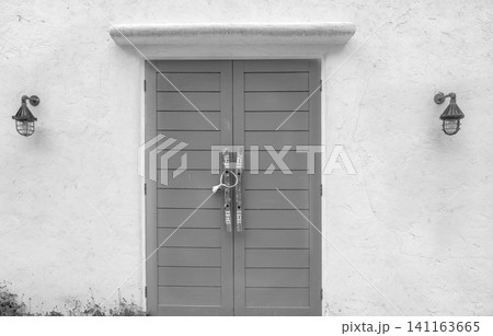 Closed wood door and white concrete wall of the building. Lamp on cement wall near the door. Building exterior. Building wall with rough texture background. Wooden door with handle at the entrance. 141163665