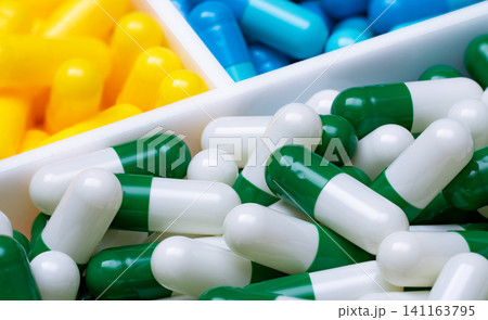 Selective focus on green-white capsule pills in plastic box on blurred blue and yellow capsule pills. Pharmaceutical industry. Pharmacy drugstore products. Background for health insurance business. 141163795