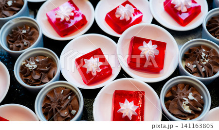 Red jelly dessert with whipped cream served on white plate and cupcakes with chocolate mousse in cup. Square red jelly plate on table at restaurant for buffet. Sweet food. Jelly agar, selective focus. Red jelly dessert with whipped cream served on white plate and cupcakes with chocolate mousse in cup. Square red jelly plate on table at restaurant for buffet. Sweet food. Jelly agar, selective focus. 141163805
