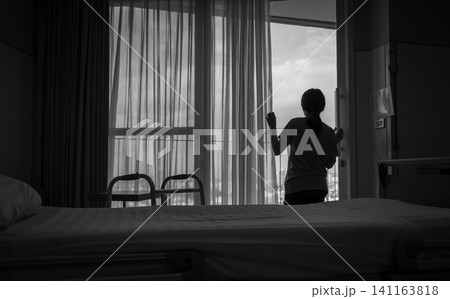 Rear view of woman holding curtain and looking out of inpatient bed room. Depressed and stressed woman. World mental health day concept. Mental health awarenes. Thoughtful woman in hospital room. Rear view of woman holding curtain and looking out of inpatient bed room. Depressed and stressed woman. World mental health day concept. Mental health awarenes. Thoughtful woman in hospital room. 141163818