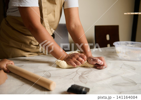 Woman kneading dough on kitchen table during home baking, hands pressing and shaping bread Woman kneading dough on kitchen table during home baking, hands pressing and shaping bread 141164084