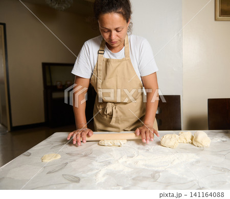 Woman rolling dough with a rolling pin on a flour-dusted table at home kitchen Woman rolling dough with a rolling pin on a flour-dusted table at home kitchen 141164088