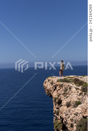 Young woman standing on edge of a high cliff looking at the deep blue sea in Malta. 141164269