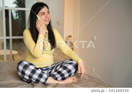 Young woman in casual attire sitting on bed having a phone conversation at home. Mobile lifestyle remote communication and home consultation Young woman in casual attire sitting on bed having a phone conversation at home. Mobile lifestyle remote communication and home consultation 141164350
