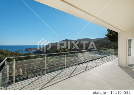 Modern balcony with glass railing overlooking coastal hills, blue sea, bright sky, sunlit terrace floor and contemporary building facade, creating calm scenic resort atmosphere Modern balcony with glass railing overlooking coastal hills, blue sea, bright sky, sunlit terrace floor and contemporary building facade, creating calm scenic resort atmosphere 141164525