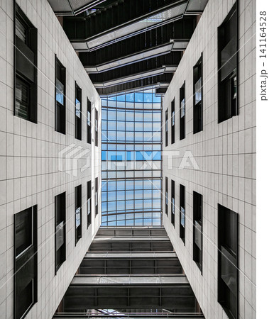 Upward architectural view through narrow atrium beneath glass skylight framed by white concrete walls, sharp geometric angles, clean lines, strong symmetry, cool daylight, modern urban structure 141164528