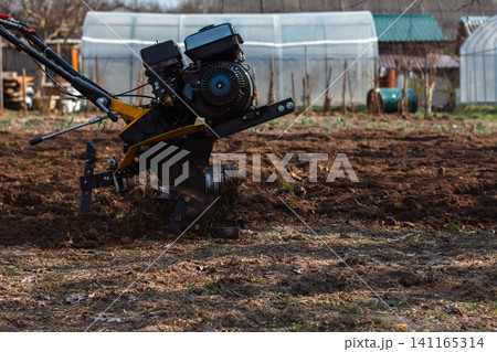 Agricultural walk-behind tractor plowing soil, preparing land for planting in a private garden with greenhouses in the background, working the earth for future crops during springtime. 141165314