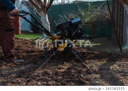 Man using a yellow walk-behind tractor to cultivate the dark brown soil in a home garden, preparing the ground for planting and showing springtime agricultural activity and gardening work. Man using a yellow walk-behind tractor to cultivate the dark brown soil in a home garden, preparing the ground for planting and showing springtime agricultural activity and gardening work. 141165315