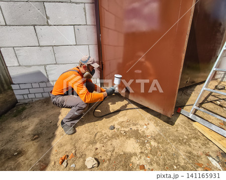 Worker wearing a respirator mask and protective clothing, using a spray gun to apply paint on a large metal surface outside a building during a renovation project. Worker wearing a respirator mask and protective clothing, using a spray gun to apply paint on a large metal surface outside a building during a renovation project. 141165931