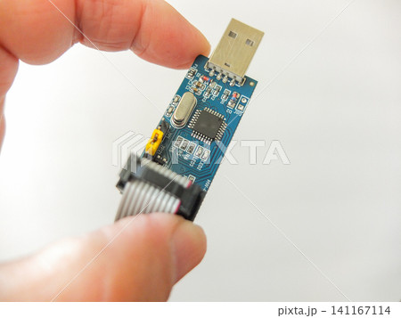 Hand holding a blue USB microcircuit electronic board programmer with ribbon cable. Hand holding a blue USB microcircuit electronic board programmer with ribbon cable. 141167114