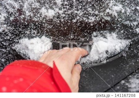 Close-up of a hand in a red sleeve reaching for a car door handle partially buried under fresh snow. Close-up of a hand in a red sleeve reaching for a car door handle partially buried under fresh snow. 141167208