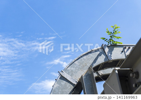 A small green plant growing resiliently out of a gap in a massive metal industrial structure under a clear blue sky. A small green plant growing resiliently out of a gap in a massive metal industrial structure under a clear blue sky. 141169963