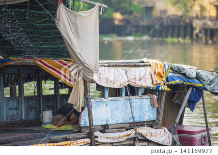 A senior man resting and sleeping inside a weathered, traditional wooden boat docked along a river canal in Vietnam. A senior man resting and sleeping inside a weathered, traditional wooden boat docked along a river canal in Vietnam. 141169977
