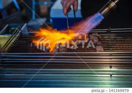 A chef using a gas blowtorch to flame-grill beef cube steaks on a wire rack at a night market. A chef using a gas blowtorch to flame-grill beef cube steaks on a wire rack at a night market. 141169979