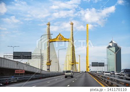 Modern city landscape with vehicles driving on the Rama IX Bridge, featuring iconic golden pylons under a blue sky in Bangkok, Thailand Modern city landscape with vehicles driving on the Rama IX Bridge, featuring iconic golden pylons under a blue sky in Bangkok, Thailand 141170110