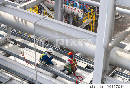 Workers wearing safety harnesses inspecting piping and steel structures at height in an industrial plant facility Workers wearing safety harnesses inspecting piping and steel structures at height in an industrial plant facility 141170144