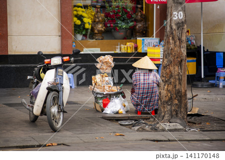 Street vendor wearing a traditional conical hat selling snacks beside a motorbike on a sidewalk in Ho Chi Minh City, Vietnam Street vendor wearing a traditional conical hat selling snacks beside a motorbike on a sidewalk in Ho Chi Minh City, Vietnam 141170148