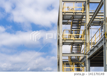 Industrial plant structure with steel stairs and yellow safety railings against a blue sky and clouds Industrial plant structure with steel stairs and yellow safety railings against a blue sky and clouds 141170149