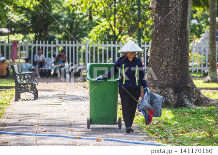 A sanitation worker wearing a traditional Vietnamese conical hat (Non La) pushing a green trash bin in a sunny park in Ho Chi Minh City A sanitation worker wearing a traditional Vietnamese conical hat (Non La) pushing a green trash bin in a sunny park in Ho Chi Minh City 141170180