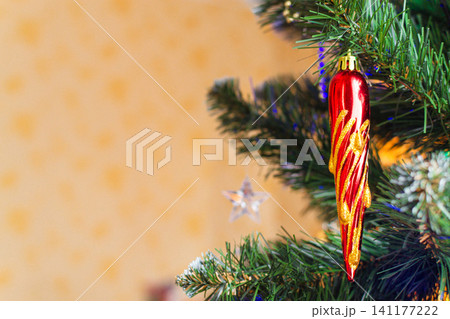 Red icicle Christmas ornament hanging on a fir tree branch with copy space. Holiday decoration and festive celebration concept for seasonal greeting and winter design. Red icicle Christmas ornament hanging on a fir tree branch with copy space. Holiday decoration and festive celebration concept for seasonal greeting and winter design. 141177222