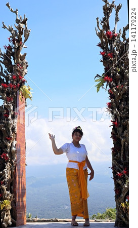 Balinese Young woman in traditional Balinese kebaya and sash posing beneath carved wood gateway with tropical foliage, smiling and raising one hand against bright blue sky Balinese Young woman in traditional Balinese kebaya and sash posing beneath carved wood gateway with tropical foliage, smiling and raising one hand against bright blue sky 141179224