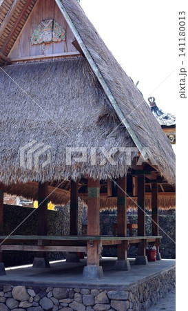 Traditional Balinese pavilion with steep thatched roof, carved wooden ornament, timber posts, raised stone base, and open shaded interior in tropical village setting Traditional Balinese pavilion with steep thatched roof, carved wooden ornament, timber posts, raised stone base, and open shaded interior in tropical village setting 141180013