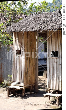 Tall bamboo huts with thatched roofs and simple wooden bench in tropical village beside weathered wall under bright sunlight and strong shadow, with bamboo, hut, huts, thatched detail Tall bamboo huts with thatched roofs and simple wooden bench in tropical village beside weathered wall under bright sunlight and strong shadow, with bamboo, hut, huts, thatched detail 141180015