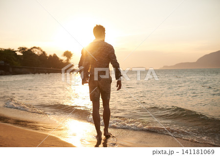 Man, walking on beach and ocean with sunset, lens flare with back view and waves on vacation in Hawaii. Tourism, travel and adventure with peace, zen and fresh air for wellness, summer and sunshine Man, walking on beach and ocean with sunset, lens flare with back view and waves on vacation in Hawaii. Tourism, travel and adventure with peace, zen and fresh air for wellness, summer and sunshine 141181069