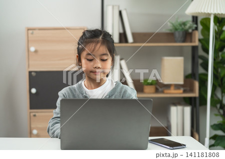 Young girl focused and working on laptop computer at desk in modern home interior with bookshelf and lamp background Young girl focused and working on laptop computer at desk in modern home interior with bookshelf and lamp background 141181808