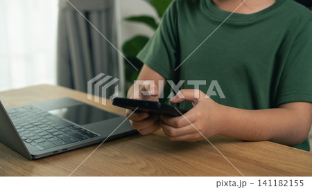 Child in green shirt holding black smartphone using device near open laptop on wooden table indoors Child in green shirt holding black smartphone using device near open laptop on wooden table indoors 141182155