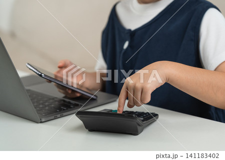 Child using calculator and holding smartphone while sitting in front of laptop working on math homework or financial calculation at white desk indoors 141183402