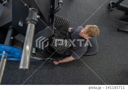 Young man performing seated leg press workout in modern commercial gym, viewed from above with weighted footplate, padded machine seat, side handles, dark clothing, and textured rubber flooring 141185711