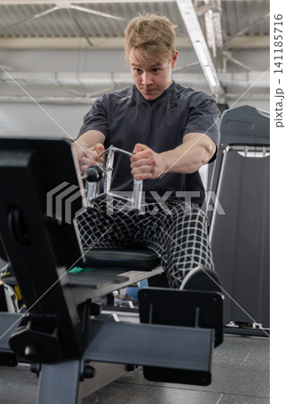 Focused young man performing intense seated rowing machine workout in modern indoor gym, gripping cable handles with feet braced on footrests under industrial ceiling lights 141185716