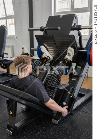 Young man performing leg press strength exercise on plate loaded machine inside bright modern gym with large windows, rubber flooring, white sneakers, and focused training posture Young man performing leg press strength exercise on plate loaded machine inside bright modern gym with large windows, rubber flooring, white sneakers, and focused training posture 141185718