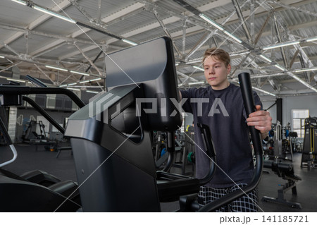 Young man exercising on elliptical trainer in spacious modern gym, holding moving handles while focused on console beneath exposed industrial ceiling with cardio and strength equipment nearby Young man exercising on elliptical trainer in spacious modern gym, holding moving handles while focused on console beneath exposed industrial ceiling with cardio and strength equipment nearby 141185721