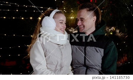 Affectionate couple sharing tender moment outdoors, wearing winter attire, standing beneath festive illumination with warm smiles and loving gaze 141185730