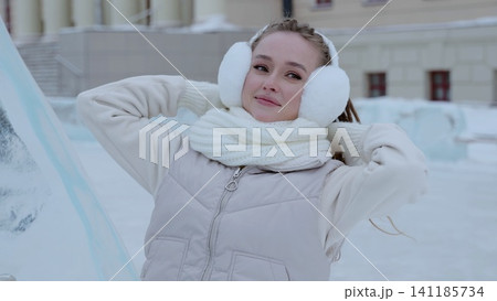 Cheerful woman bundled in earmuffs and scarf relaxing outdoors, leaning back with hands behind head near intricate winter ice sculptures 141185734
