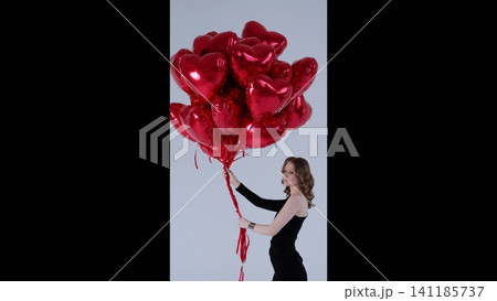 Elegant woman in black dress posing with red heart balloons, conveying romantic mood in minimalist studio setting against white backdrop Elegant woman in black dress posing with red heart balloons, conveying romantic mood in minimalist studio setting against white backdrop 141185737