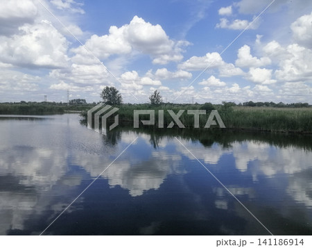 Reflection of white clouds on calm river forms natural harmony. blue sky is reflected in calm waters of nature small pond. Reflection beauty and peace calm feeling to water composition. Reflection of white clouds on calm river forms natural harmony. blue sky is reflected in calm waters of nature small pond. Reflection beauty and peace calm feeling to water composition. 141186914