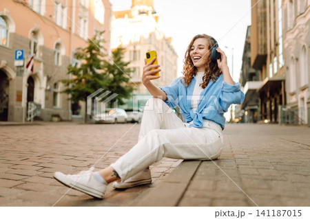 Happy relaxed woman in wireless headphones choosing, listening favorite music in urban city street. 141187015