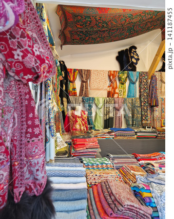 Paris, France - December 10, 2025: vibrant market stall is filled with colorful, patterned scarves, shawls, and textiles. Stacks of folded fabrics of rich, intricate craftsmanship rest on the counter 141187455