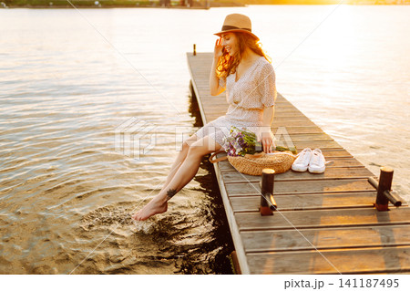 Young woman in dress sitting on wooden pier near pond. Rustic style of wedding. Travel, tourism 141187495