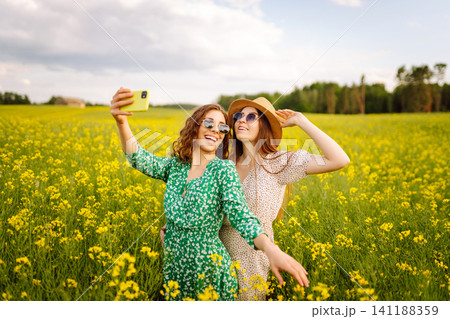 Two Young woman takes a selfie while walking flowering field. Relax and lifestyle. Summer landscape. 141188359