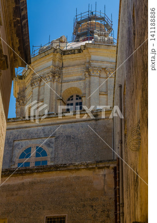 Baroque limestone cathedral dome with scaffolding seen between narrow buildings against blue sky in Mdina, Malta Baroque limestone cathedral dome with scaffolding seen between narrow buildings against blue sky in Mdina, Malta 141188886