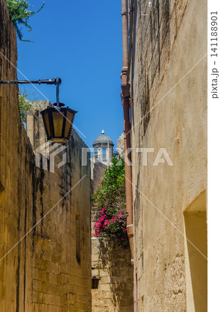 Narrow medieval alley with old lantern, pink bougainvillea and church dome in golden limestone street in Mdina, Malta 141188901
