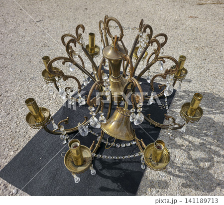 A vintage brass chandelier with crystal glass pendants is displayed on a black mat at a flea market. The sun shines down as people browse nearby stalls looking for treasures. 141189713
