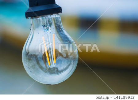 Closeup of a hanging outdoors incandescent glass lamp with glowing filaments. Vintage style light bulb. Copy space. Blurred background. energy, innovation, electricity, sustainability, illumination Closeup of a hanging outdoors incandescent glass lamp with glowing filaments. Vintage style light bulb. Copy space. Blurred background. energy, innovation, electricity, sustainability, illumination 141189912