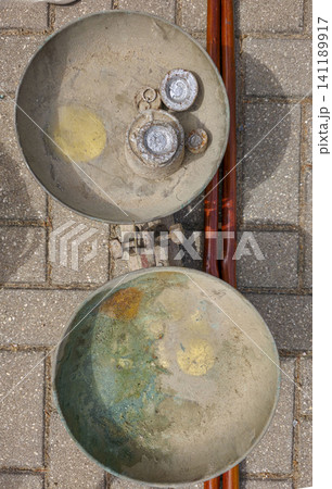 An antique balance scale with a rusty appearance. Old metal weights rest inside the scale on a brick pavement. The bricks show signs of age and wear. An antique balance scale with a rusty appearance. Old metal weights rest inside the scale on a brick pavement. The bricks show signs of age and wear. 141189917